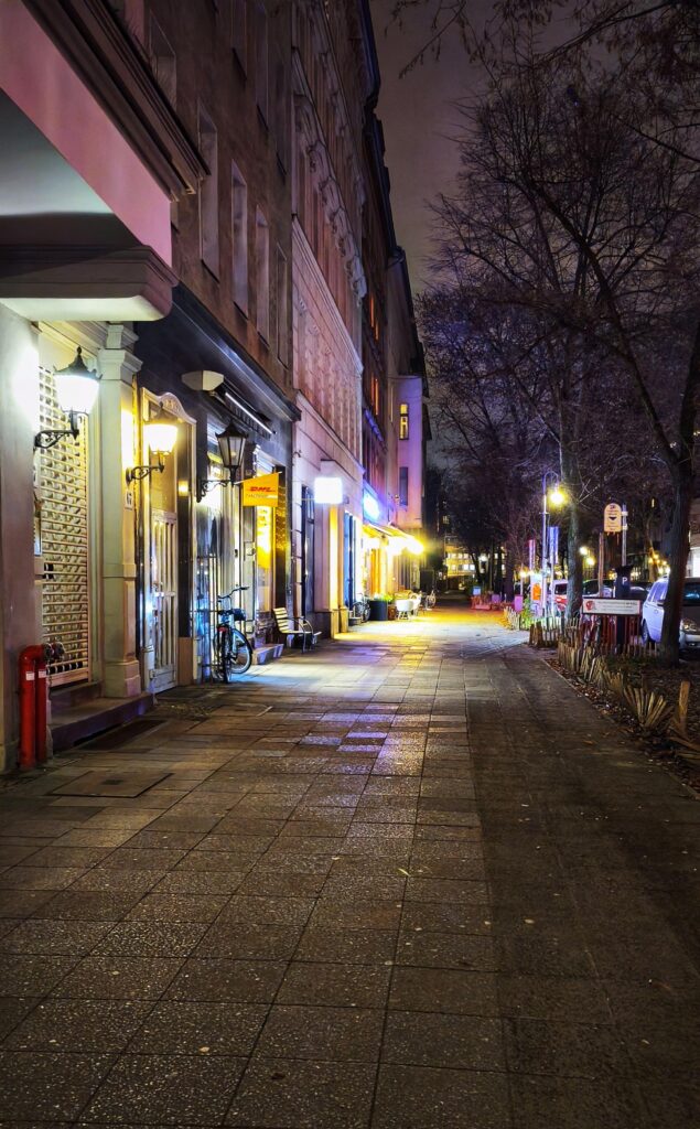 Nachtaufnahme, Häuser uns Strasse relativ hell beleuchtet durch die Neonlichter bei Geschäften und Kneipen, gelblich und violett überwiegt: Man sieht links die Häuserzeile, in er Mitte den Gehsteig, mit Steinplatten geebnet und rechts teilweise Bäume, die die Szenerie begrenzen
